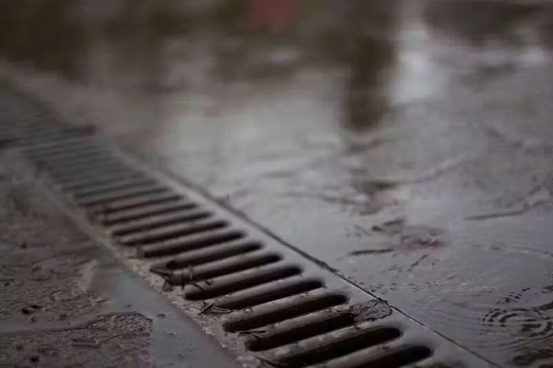 Grelha de ralo no chão molhado, com água escorrendo e gotas de chuva formando círculos