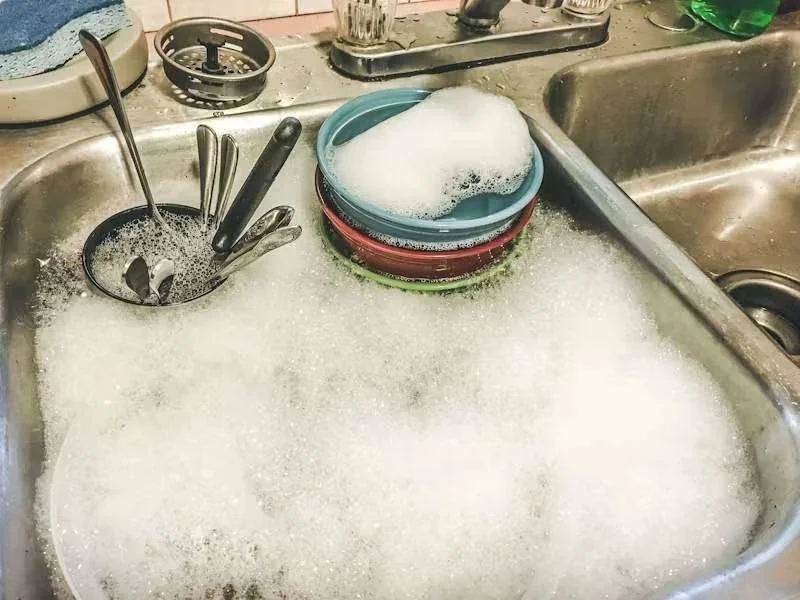 Pia de cozinha de inox com água e muita espuma, cheia de louça suja, talheres e tigelas empilhadas