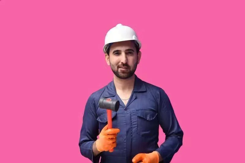 Homem sorrindo, de capacete branco e uniforme azul, segurando martelo de borracha com luvas laranja