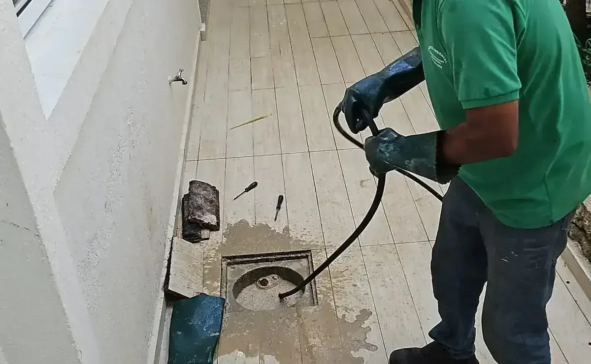 Profissional com luvas desentupindo ralo no chão com cabo, ferramentas e água ao redor