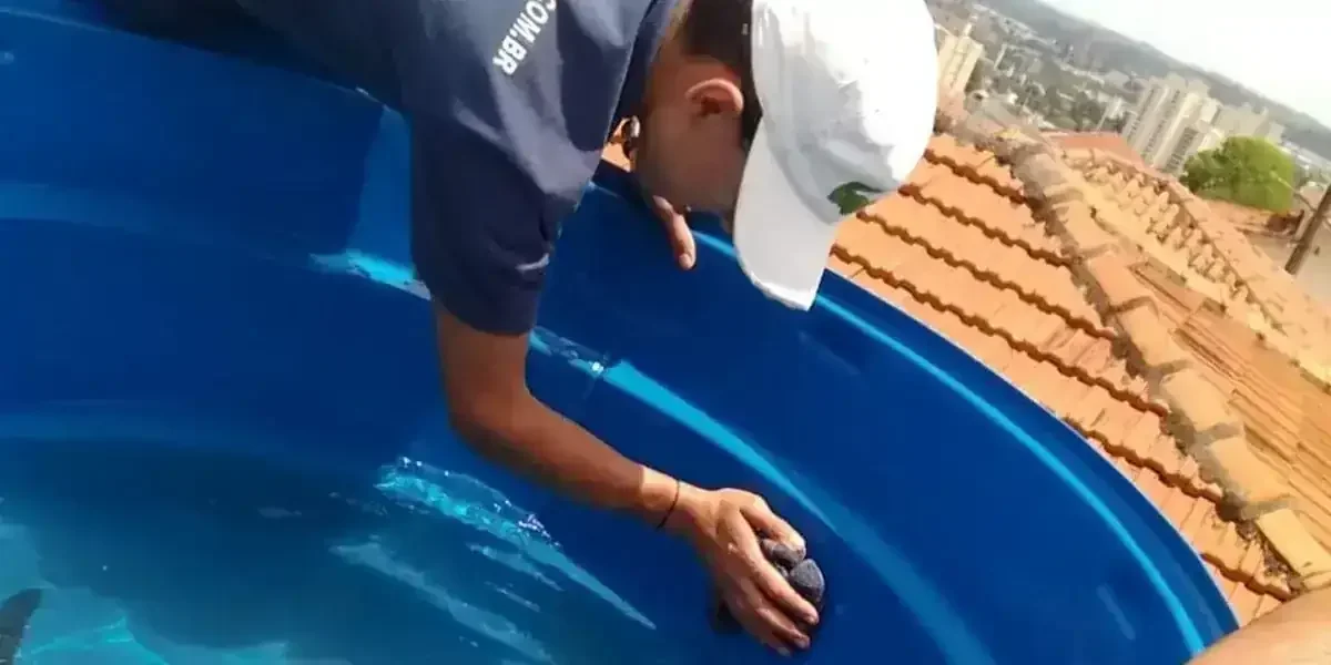 Pessoa de boné branco limpando o interior de um tanque azul, com telhado de cerâmica e prédios ao fundo