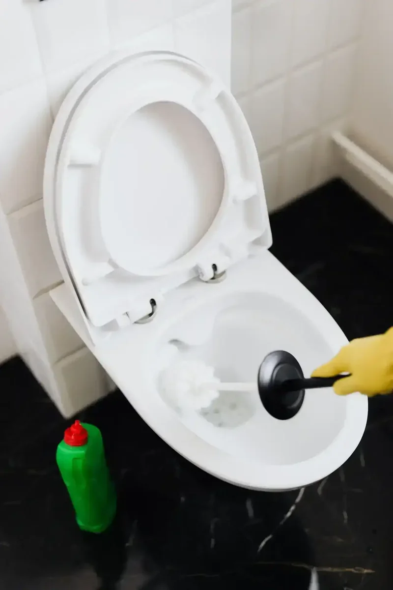 Mão com luva amarela limpando vaso sanitário com escova, produto de limpeza verde no chão