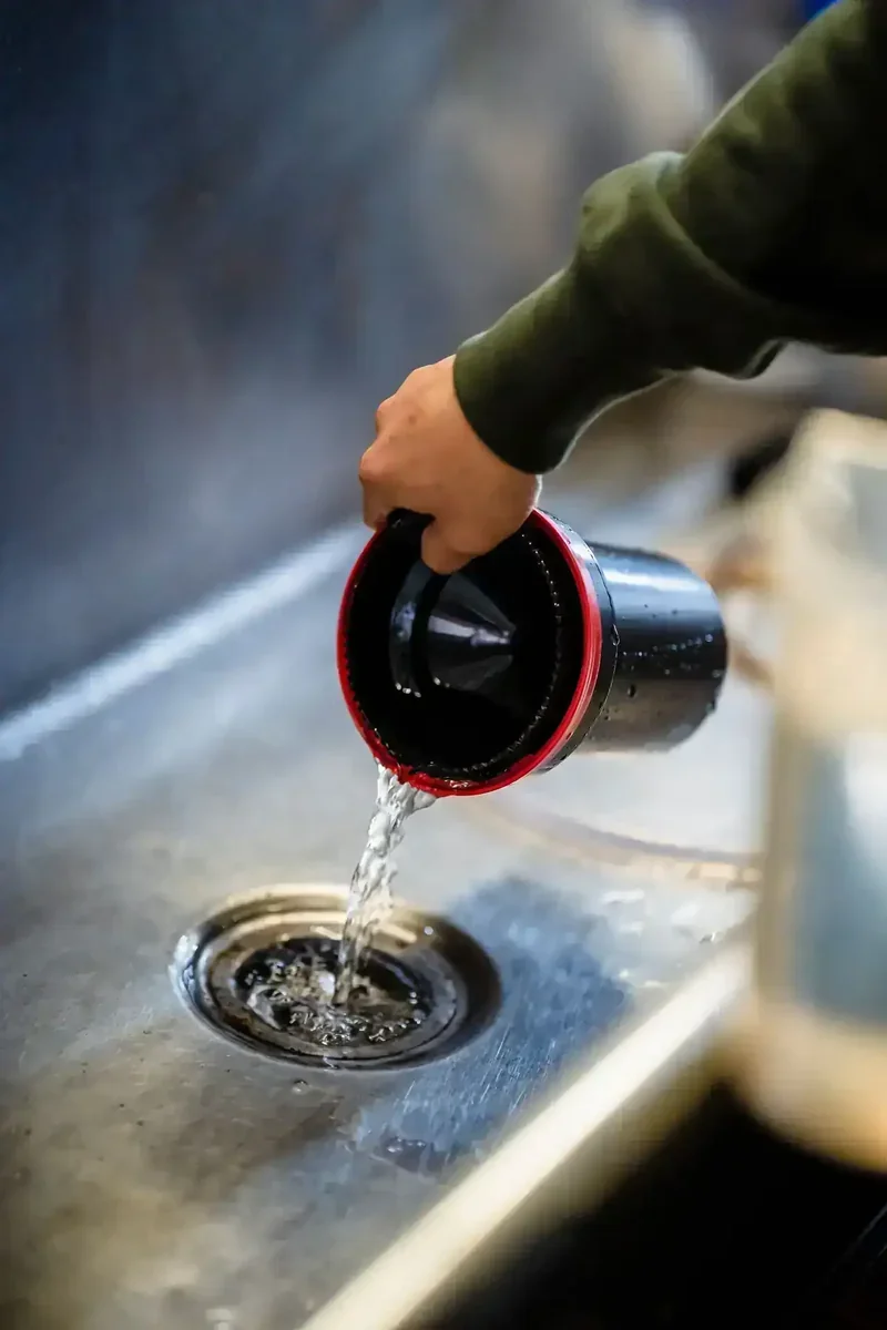 Mão derramando água de recipiente preto com borda vermelha em ralo de pia de inox