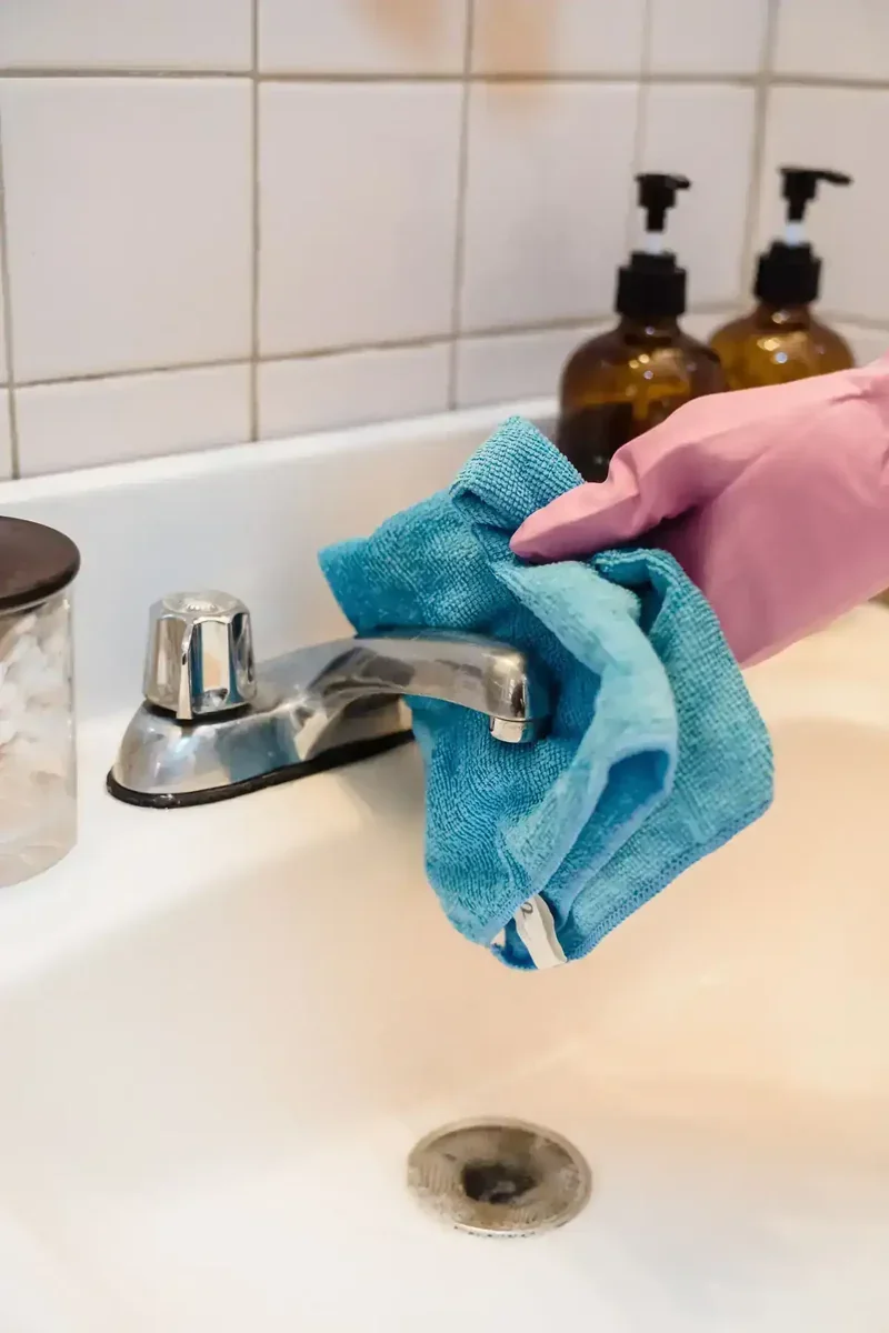 Mão com luva rosa limpando torneira de pia de banheiro com pano azul. Azulejos brancos e frascos marrons ao fundo